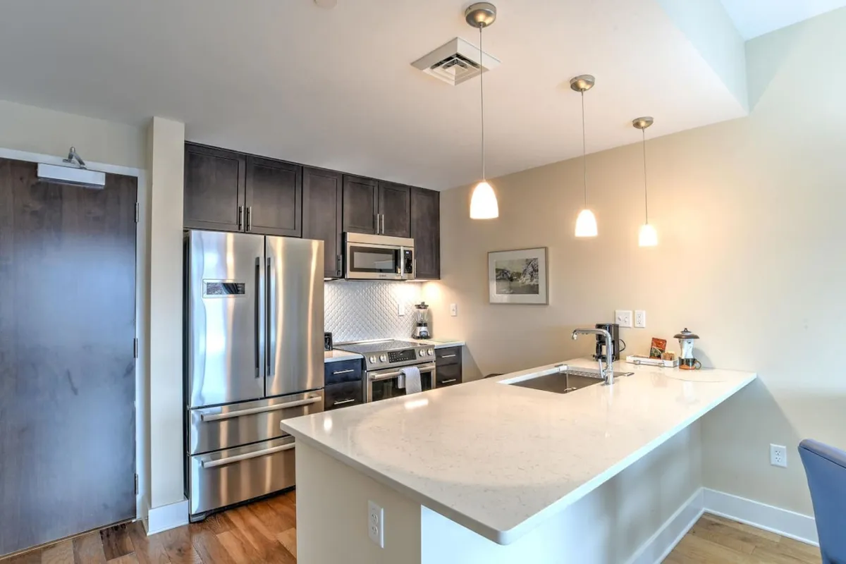 A wide-angle view of the kitchen, highlighting white countertops, a kitchen island, and pendant lights, with stainless steel appliances in the background. The setup is warm and inviting, ideal for group meals or family gatherings.