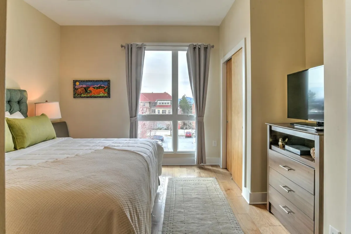 A contemporary bedroom highlighted by a large window letting in natural light, a comfortable bed with crisp linens, tasteful artwork, and a flat-screen TV. The atmosphere is calm and inviting, suggesting a restful retreat after a busy workday or a quiet spot to catch up on emails.