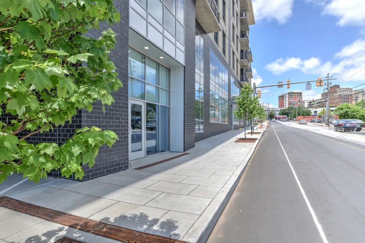 This exterior image features a modern building facade with large glass windows, surrounded by sidewalk greenery. The scene shows people walking and biking, capturing the lively, walkable atmosphere of a downtown area. The presence of greenery and street life evokes the vibrancy and accessibility described in Asheville's downtown.
