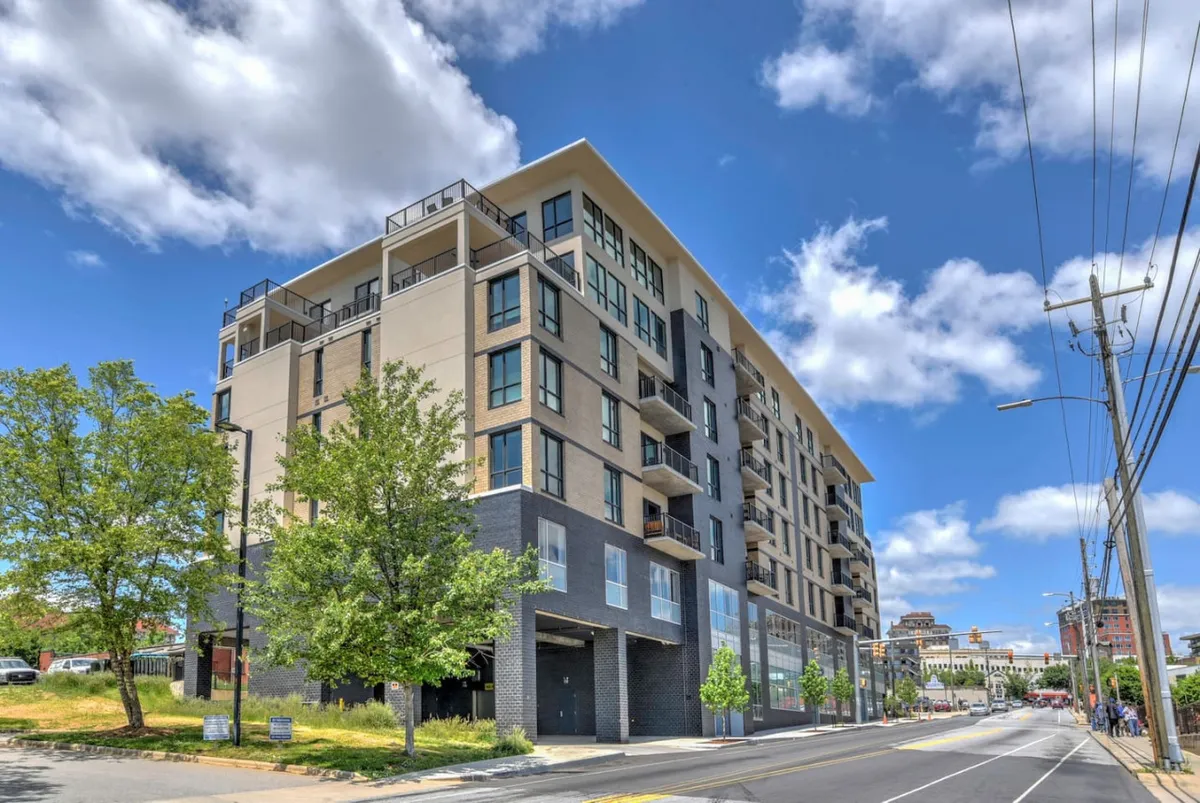 A wider exterior perspective shows the modern apartment building surrounded by lush greenery and a lively street view. The vibrant urban setting emphasizes both convenience and a sense of community as guests arrive.