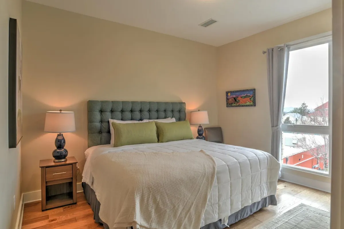 This image showcases a serene bedroom with a plush bed featuring a tufted headboard, a nightstand with a lamp, tasteful artwork on the wall, and a large window allowing natural light to fill the space. The room exudes a sense of calm and comfort, ideal for restful sleep and quiet moments.
