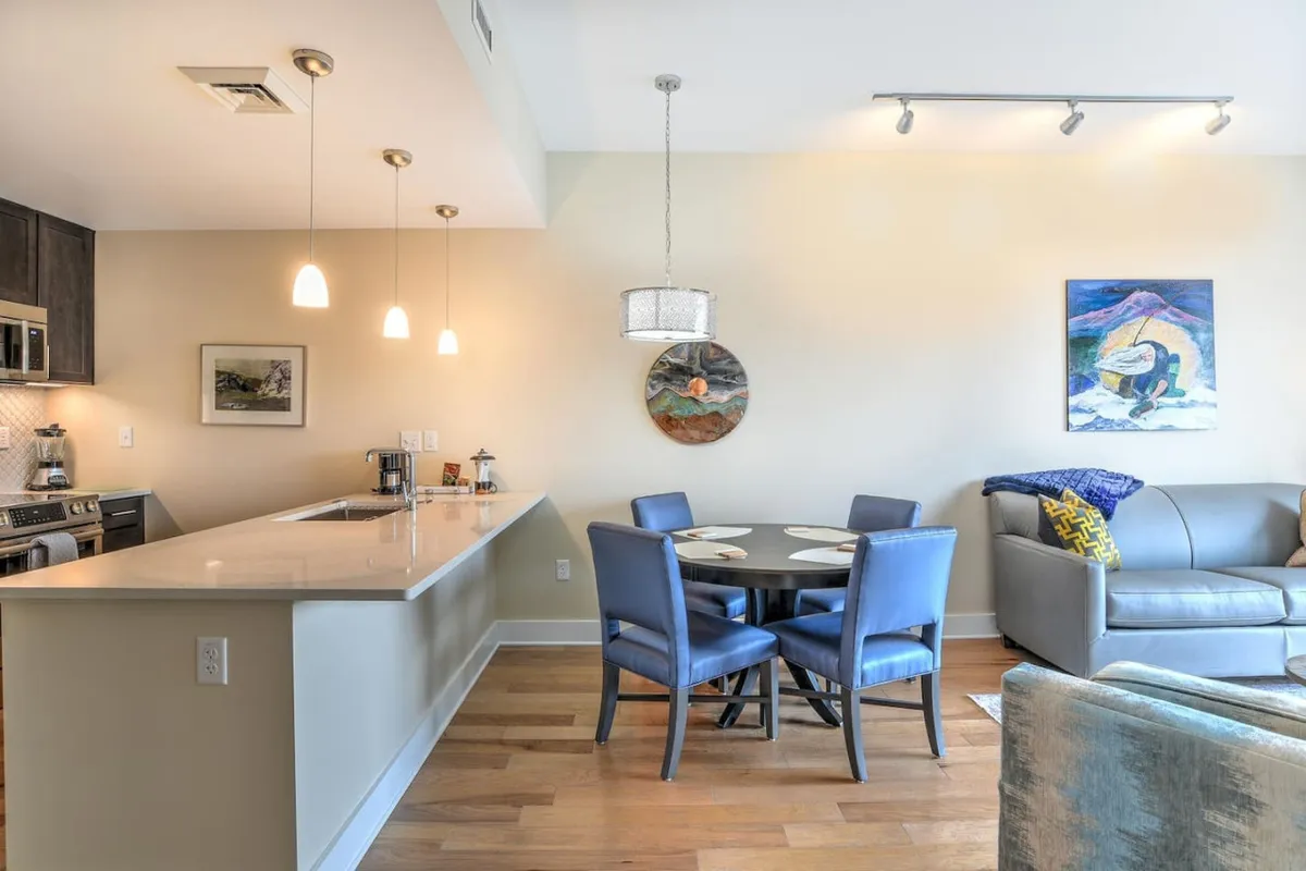 A wide view of the living and dining area, showing a modern sofa, a kitchen island, elegant artwork, and a dining table. The composition emphasizes a contemporary yet comfortable mood, ideal for both relaxing and spending time with friends or family.